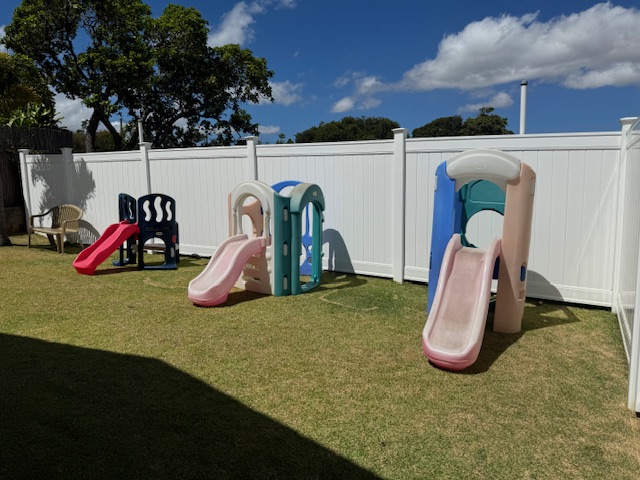 Playground area with two small slides and seating, enclosed by a white fence, under a blue sky.