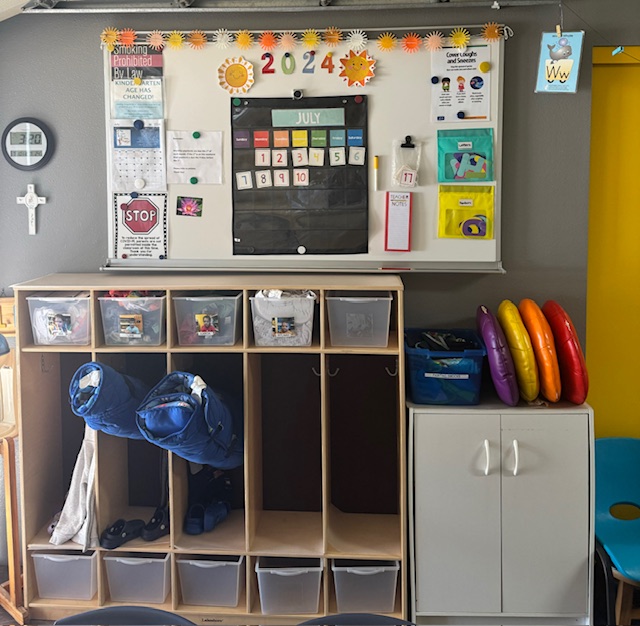 A classroom storage area featuring a calendar, educational materials, and colorful trays for children's belongings.