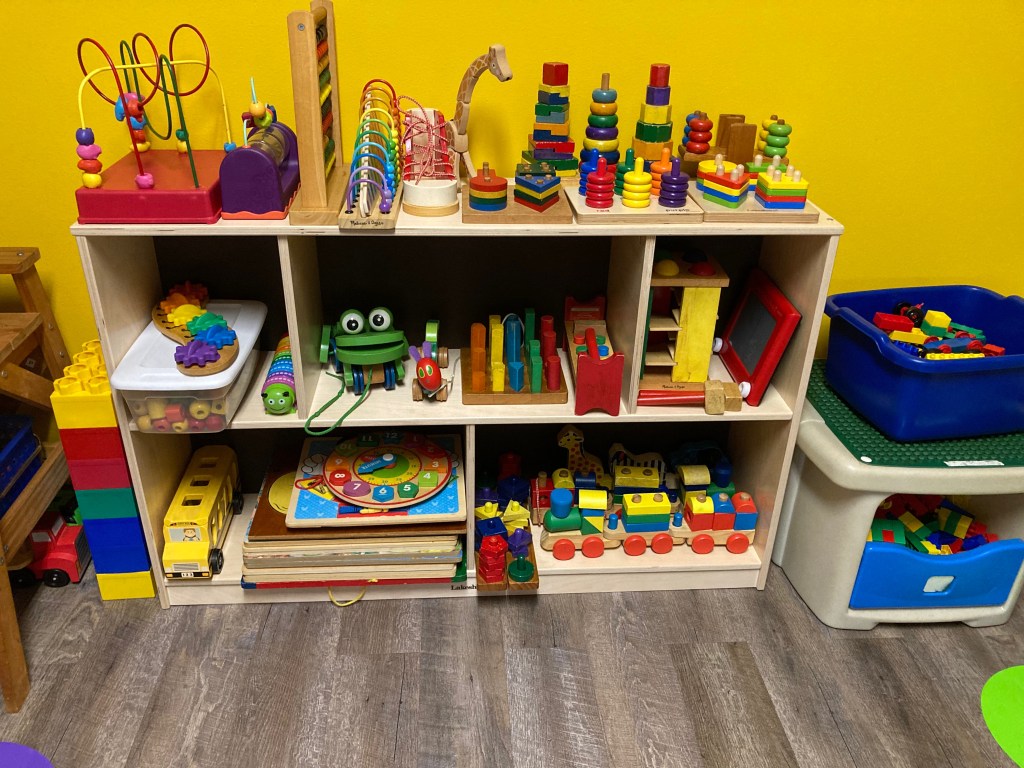 A colorful shelf filled with various educational toys and games for children, including stacking rings, a frog toy, a train set, and a collection of building blocks, all set against a bright yellow wall.
